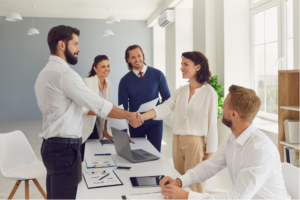 Two persons shaking hands in a meeting among other people.