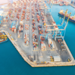A zoomed-out photo of a port with cargo on the ground and ships around.